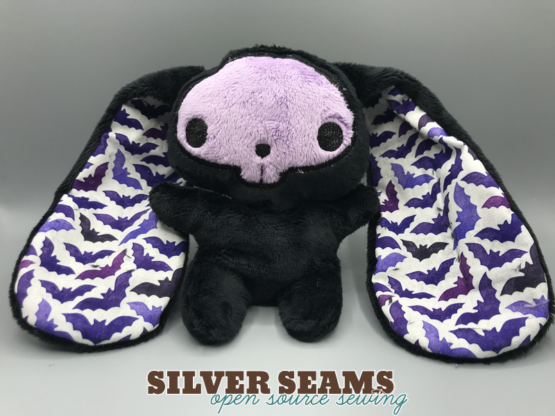 A black plush bunny with a lavender applique kawaii skull on its face, and watercolor purple-on-white bat fabric lining its ears, which are very long and wide.