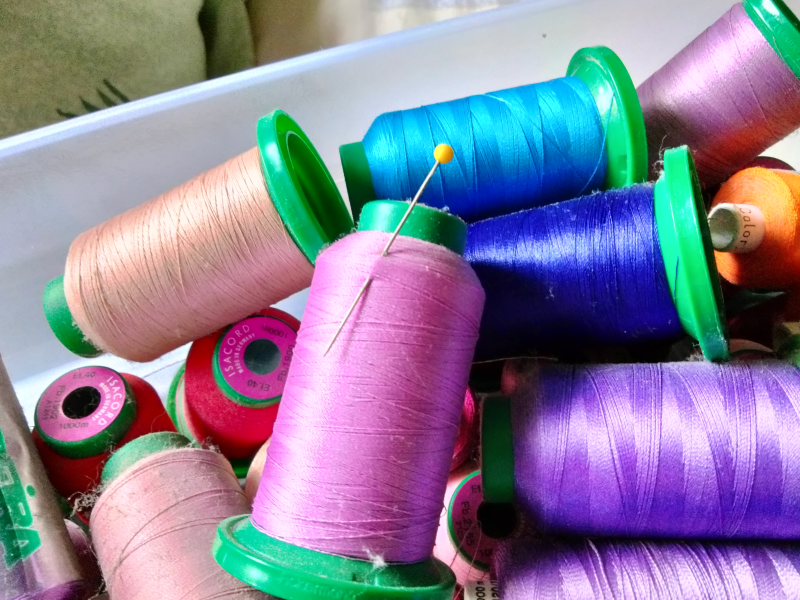 A box of dusty thread cones, including one with a long pin stuck through the shoulder of the thread