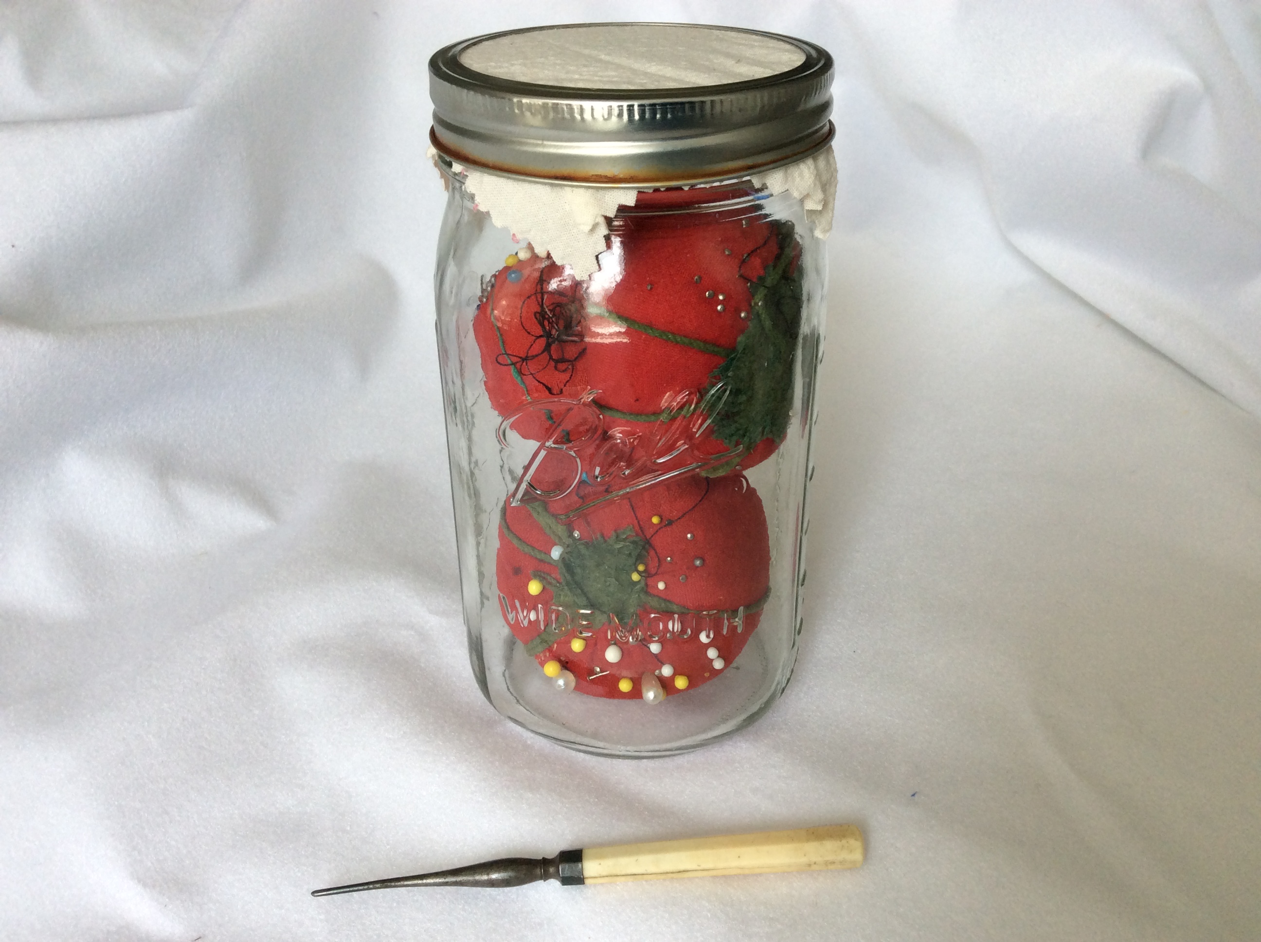 A canning jar containing tomato-shaped pincushions, and an antique tailor's awl.